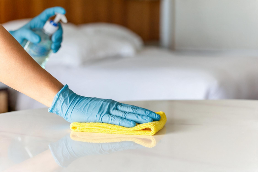 Mão com luva a limpar a superfície de um quarto de hotel com pano de microfibra amarelo e pulverizador